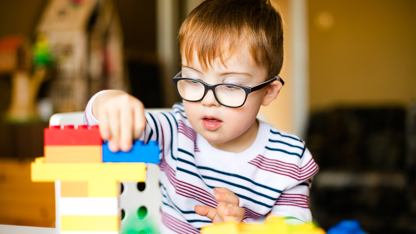A boy with red hair and a striped shirt plays with large Lego blocks. 