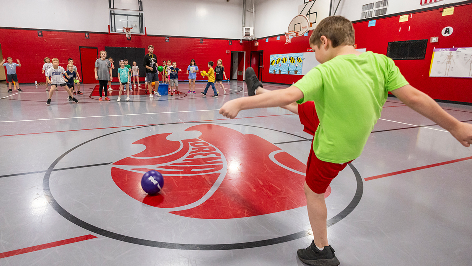 A young child, wearing a green shirt and red shorts, kicks a ball to his peers within a gymnasium. 