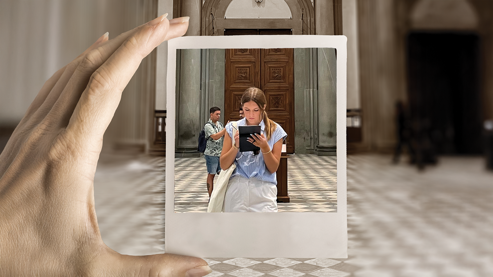 A hand holds a Polaroid photo of a young woman writing on a tablet while inside a museum.