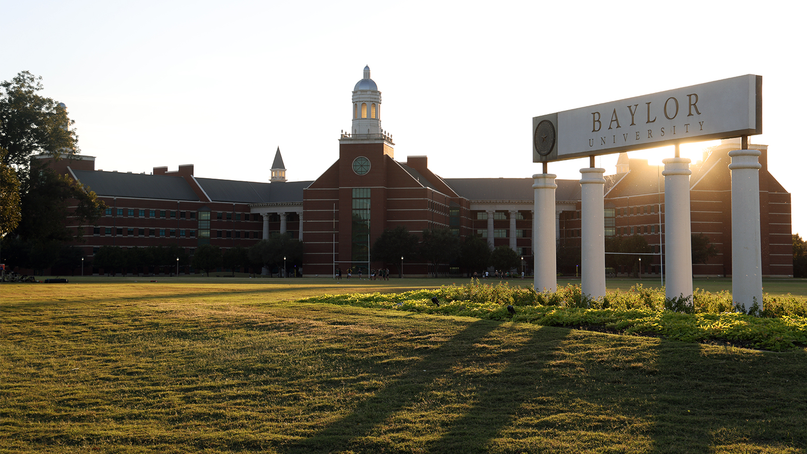 As the sun sets, light shines on Baylor's sign with four white pillars and campus building in background.