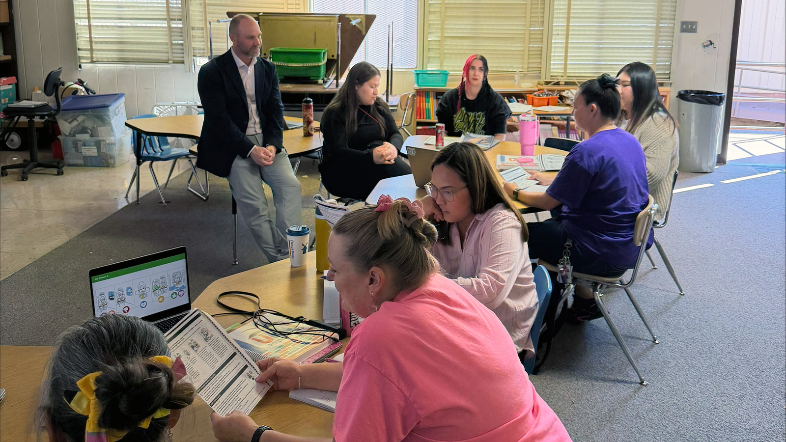 Teachers use reading materials and electronics, while sitting at two classroom tables