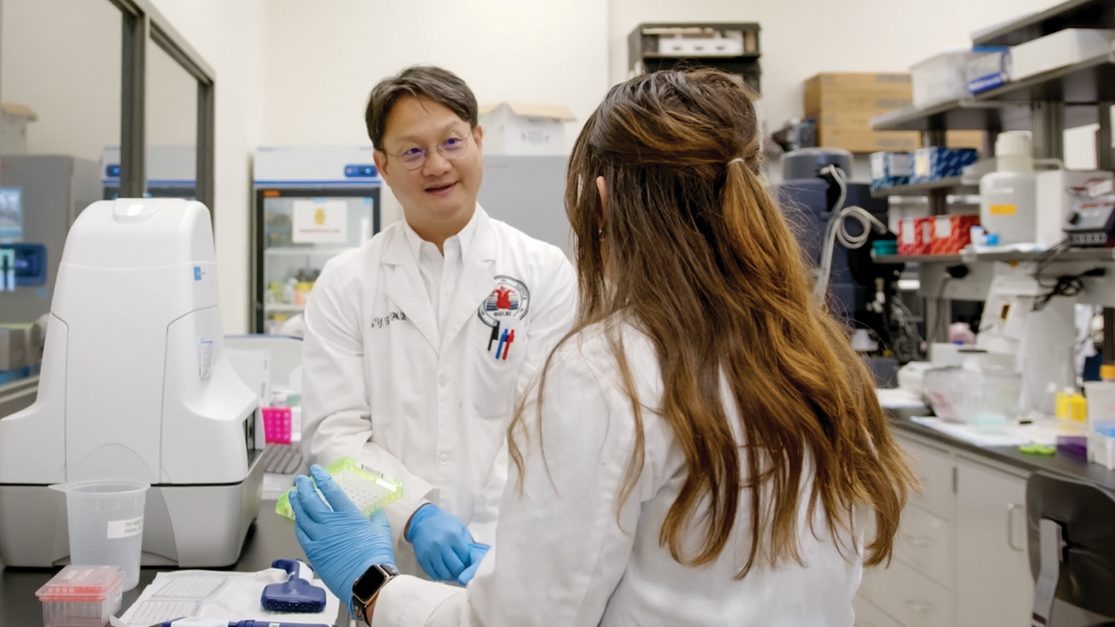 Two people, wearing white lab coats, speak to each other while working with lab equipment.