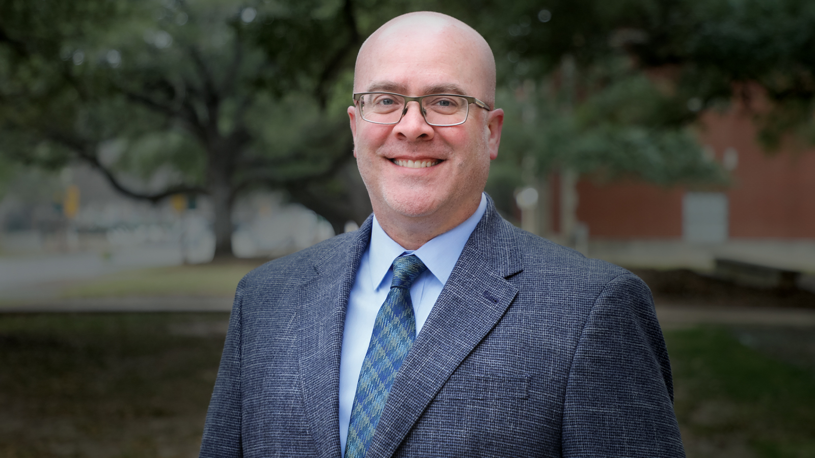 Matt Brosi, wearing a suit jacket and tie, standing with trees in the background