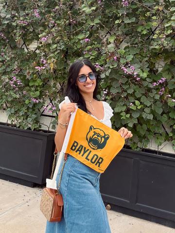 Paola Medina López holds a yellow Baylor University flag, while standing in front of a wall of flowers.