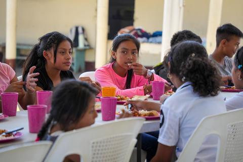 Children sit at a communal table and eat a nutritious meal.