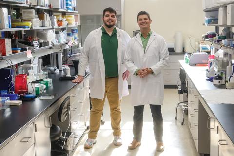 Two men, wearing white lab coats, stand in an isle with lab equipment on both sides.