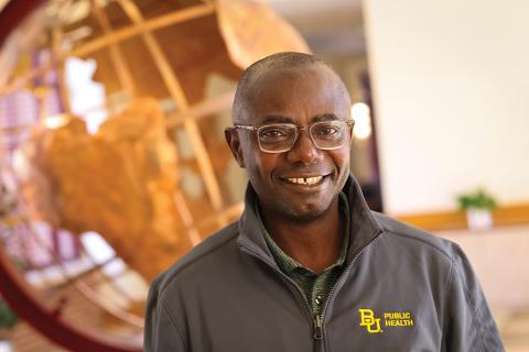 Matt Asare wears a grey "BU Public Health" sweater while standing in front of a gold globe.