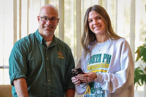 Man with green button up shirt stands next to woman with grey sweatshirt holding cell phone