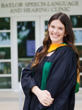 Bethany Neubert, dressed in her graduation gown stands in front of the Baylor Speech-Language and Hearing Clinic.