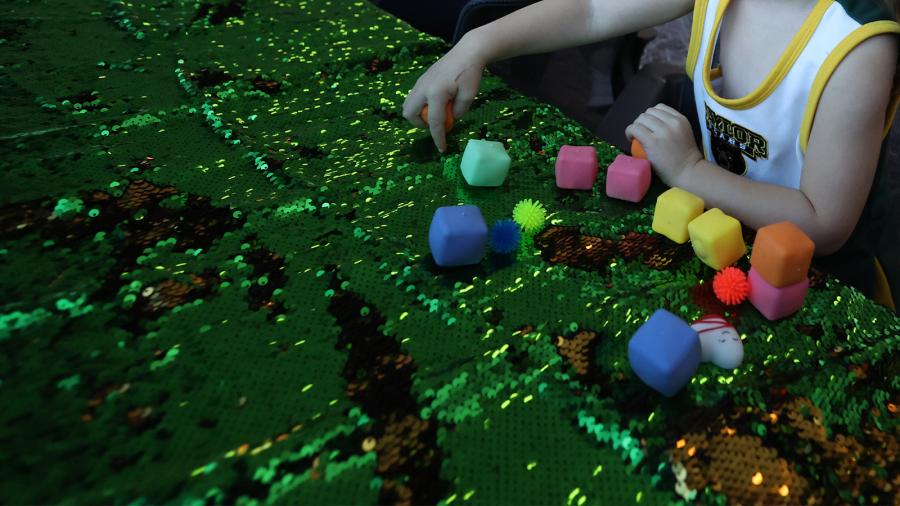 A child plays with sensory items like squishy shapes and a sequin tablecloth. 
