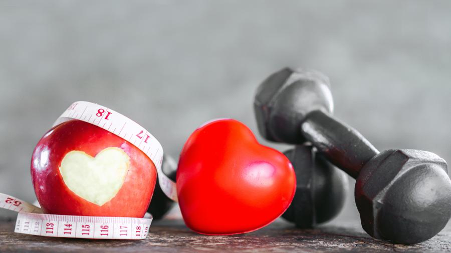 An apple with a heart space cut into it, a tape measure, and black hand weights against a grey background.