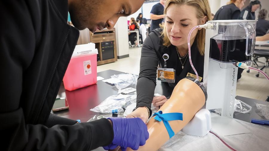 Physician Assistant faculty member walks a male student through the process of administering an IV on a high-fidelity manikin arm.