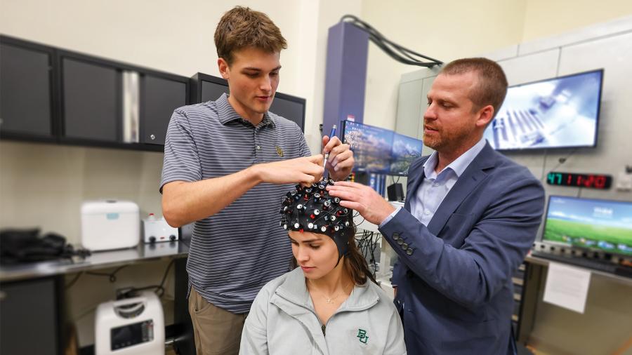 Woman wearing grey sweatshirt and a research cap to test the brain sits, while two men adjust the placement of cap.