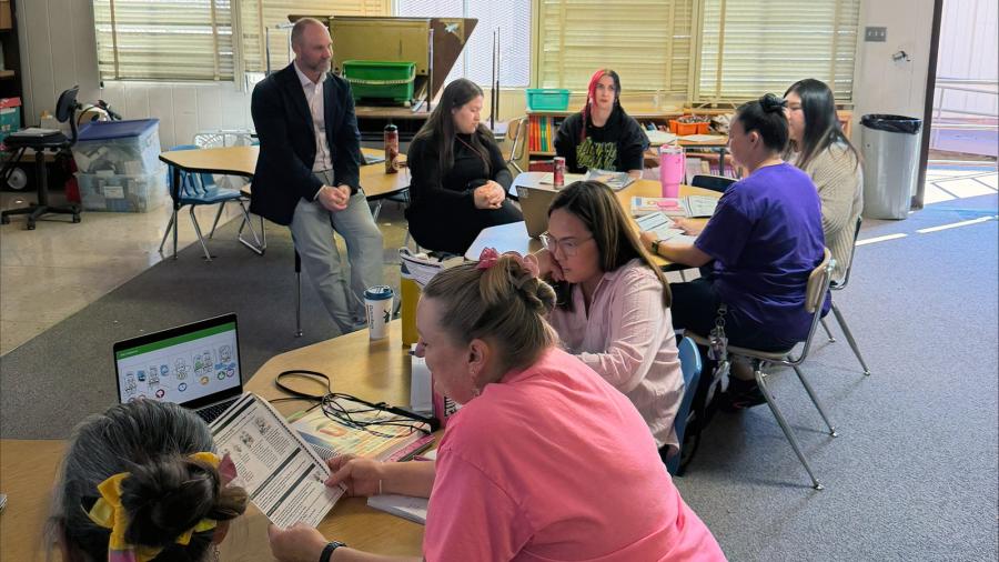 Teachers use reading materials and electronics, while sitting at two classroom tables