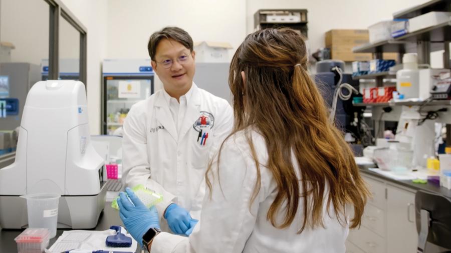 Two people, wearing white lab coats, speak to each other while working with lab equipment.