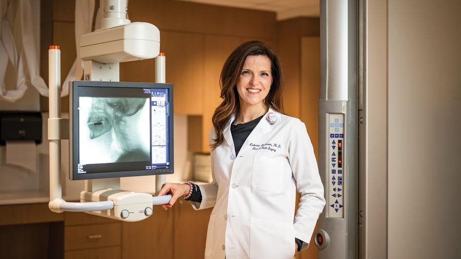 Katherine A. Hutcheson, in a white lab coat, stands next to an X-ray machine that images a human head and neck.