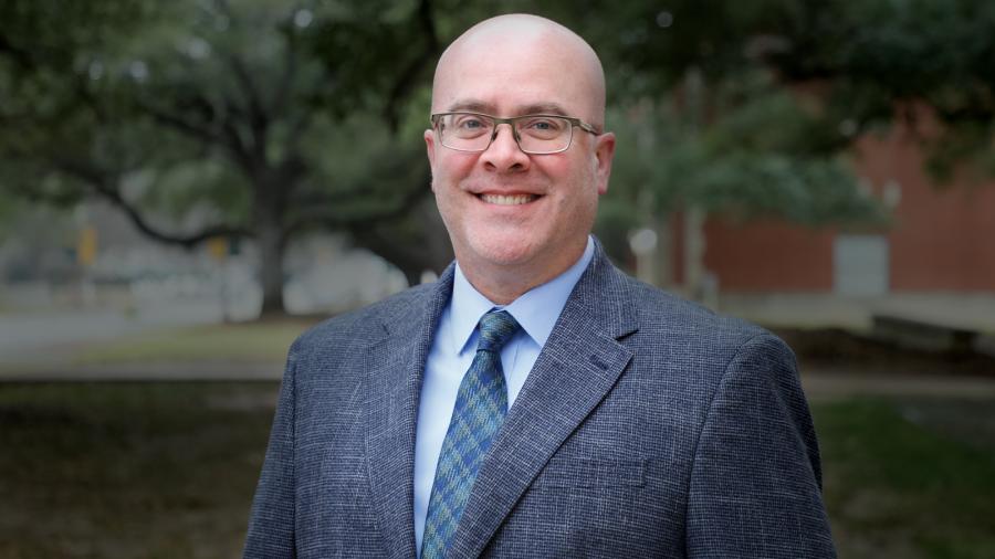 Matt Brosi, wearing a suit jacket and tie, standing with trees in the background