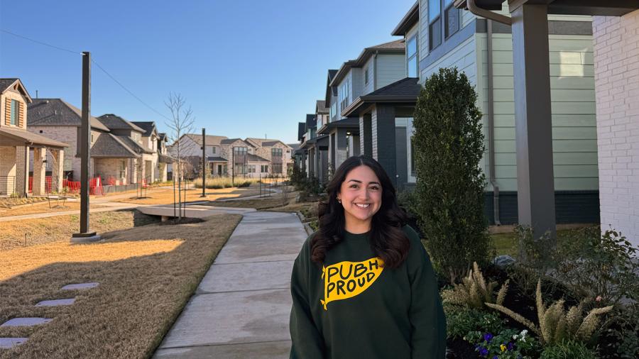 Serena Enriquez, wearing a Baylor University Public Health sweatshirt, stands within a newly built agrihood community's row of homes.