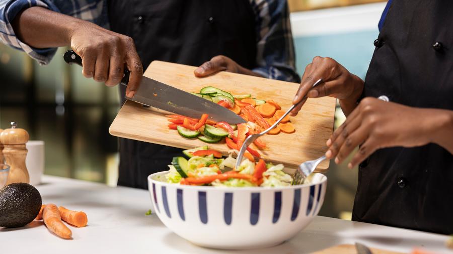 Two people chop vegetables and add them to a salad.